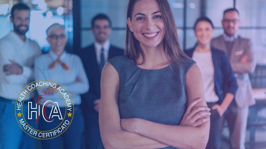 Donna sorridente in primo piano con un gruppo di colleghi sullo sfondo e logo Health Coaching Academy, simbolo di leadership e collaborazione.